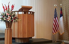 From left to right: flower arrangement, pulpit, U.S. flag, and the Christian flag on flag poles with eagle and cross finials respectively, in Covenant Presbyterian Church, Long Beach, California, United States.