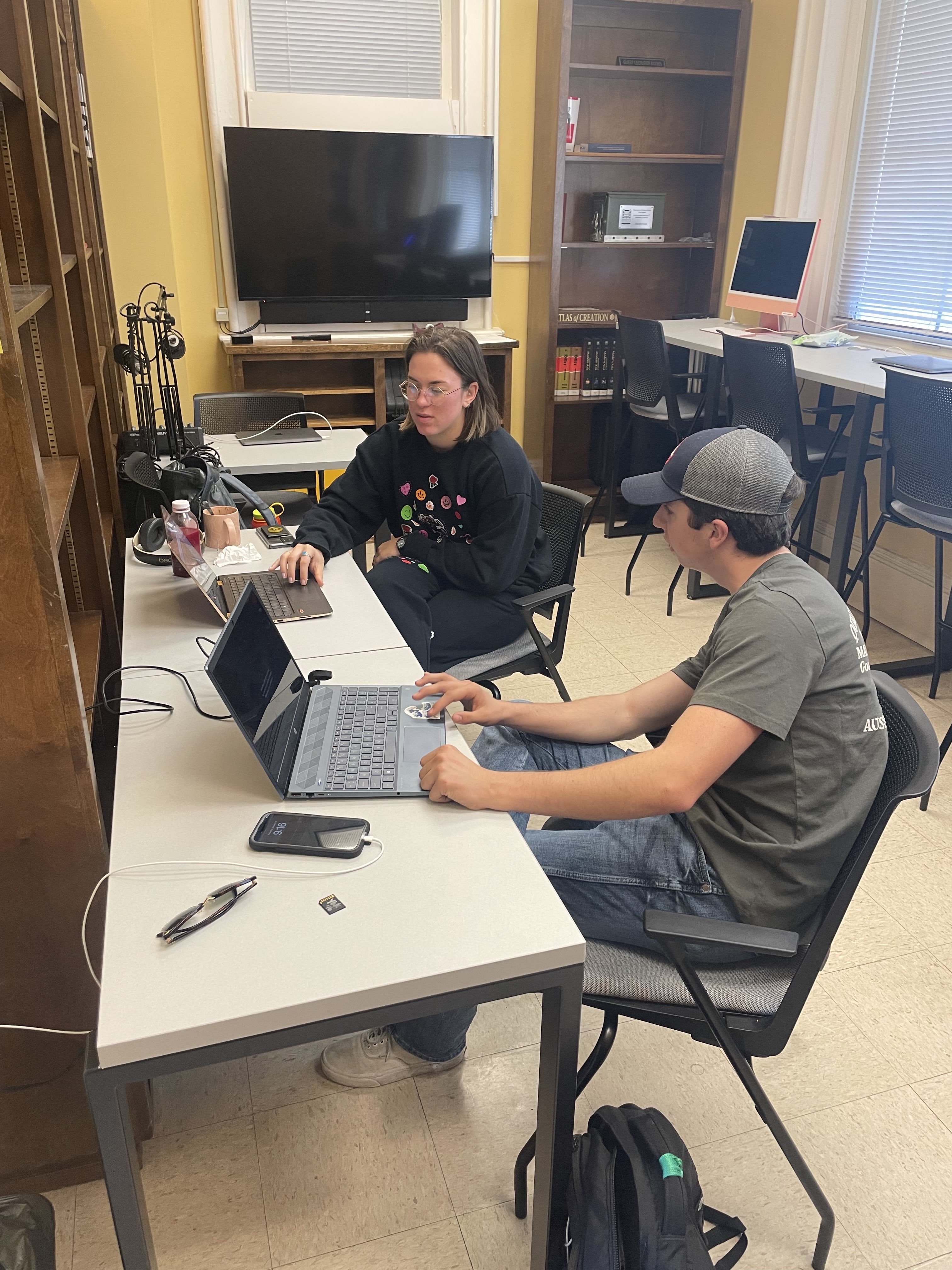 Two students are working on a podcast next to a microphone, laptop, and recording equipment.