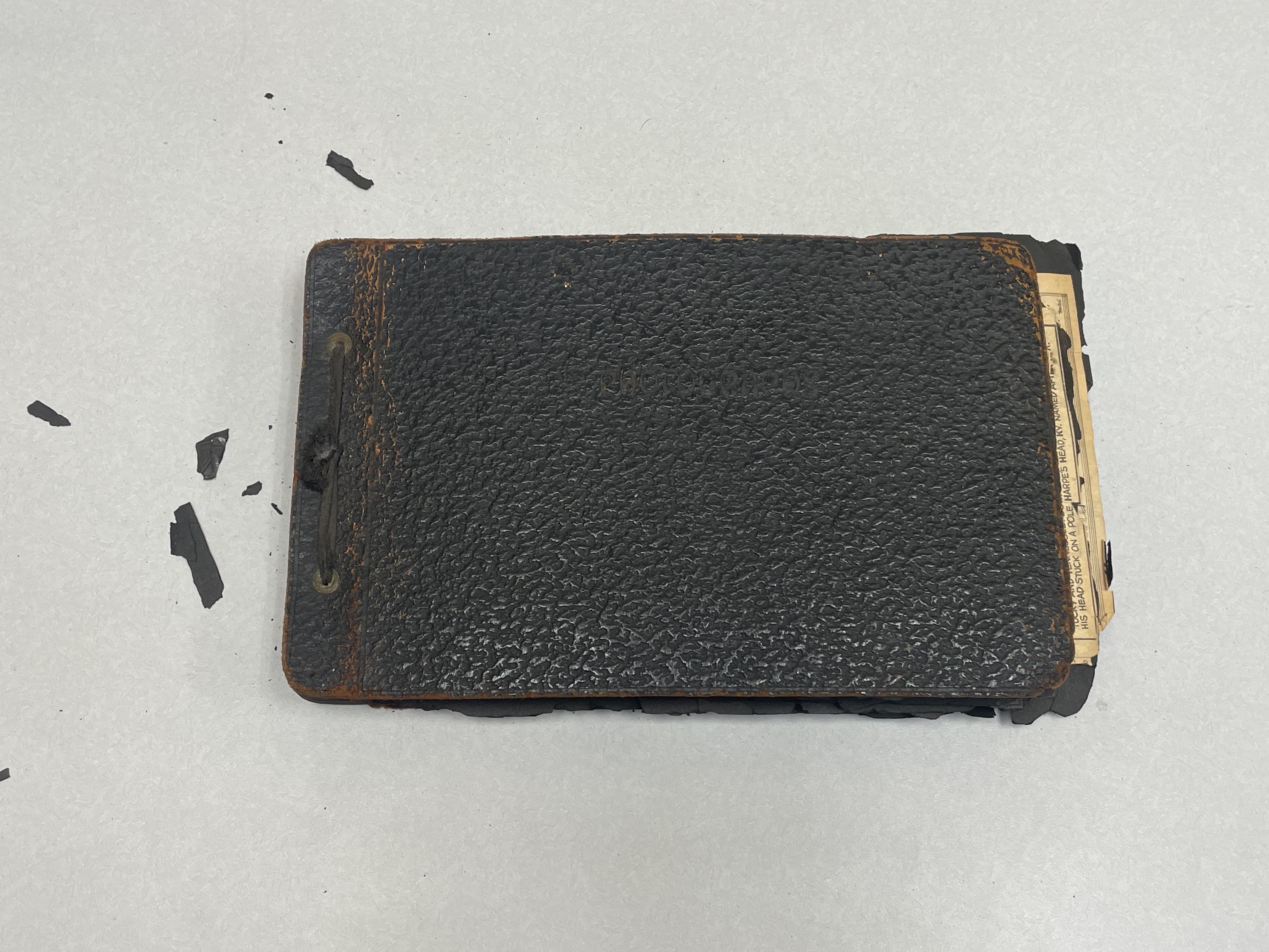 A dilapidated scrapbook. There are newspaper clippings sticking out from the edge of the landsscape-oriented book. There are also pieces of black paper paper falling off the cover.