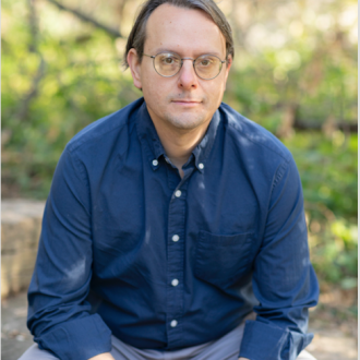 Picture of James Bielo. He's wearing a blue, button-down shirt and grey pants. He wears glasses with round frames and looks into the camera. In the background is a blur of stone pavements and foliage.