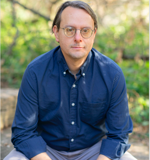 Picture of James Bielo. He's wearing a blue, button-down shirt and grey pants. He wears glasses with round frames and looks into the camera. In the background is a blur of stone pavements and foliage.